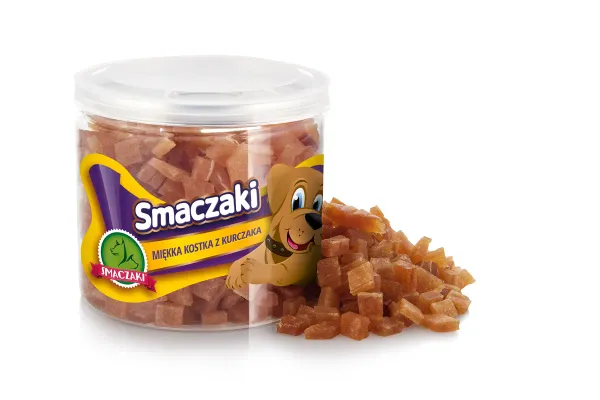 Przezroczysty słoik z żółtą etykietą marki stoi na białym tle, wypełniony drobnymi pomarańczowymi kostkami, a garść tego samego towaru leży rozsypana obok.
